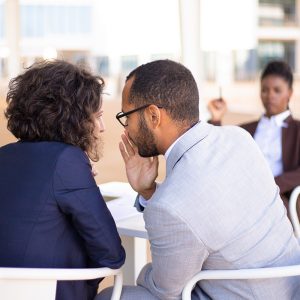 Employees gossiping about young female colleague. Business man and woman whispering, African American employee sitting in background. Office rumors concept